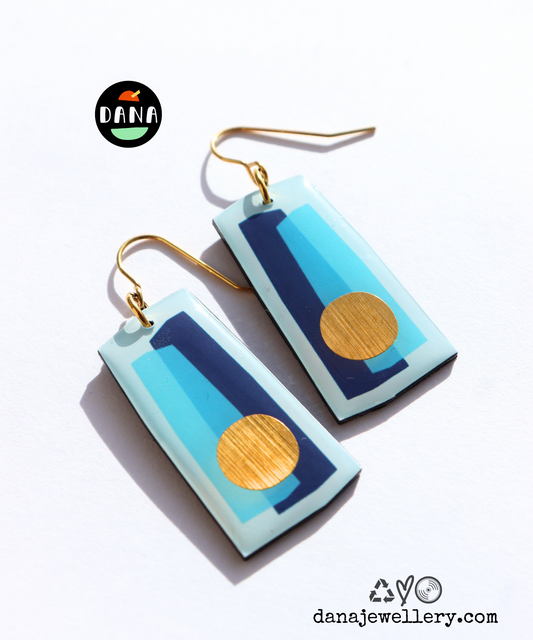A pair of drop earrings handmade in Ireland featuring abstract geometric patterns in blue shades with a gold circle detail, made from recycled vinyl records, with gold-plated hypoallergenic hooks.