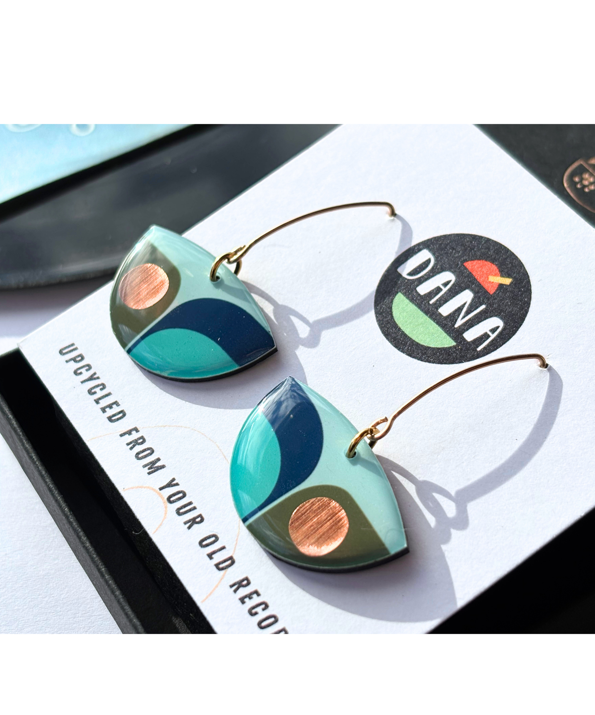 A pair of retro inspired drop earrings featuring geometric patterns in blue, green, and teal colours with copper accents with long stylish hooks, made in Ireland from recycled vinyl record, displayed on a DANA Jewellery branded card.