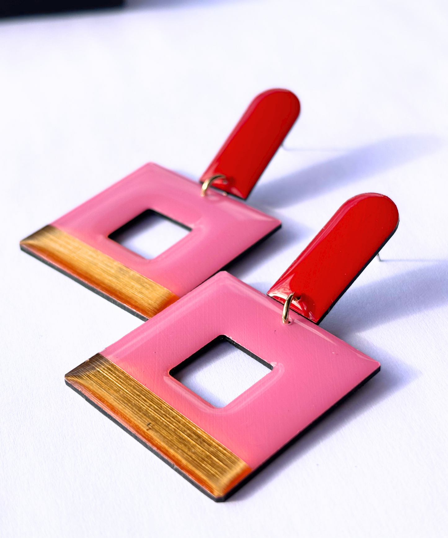 A pair of statement geometric earrings with bright red long arch posts and dangly square contours in pink with a shiny gold band running along the base, against a white background. Handmade in ireland from recycled vinyl record by DANA Jewellery.