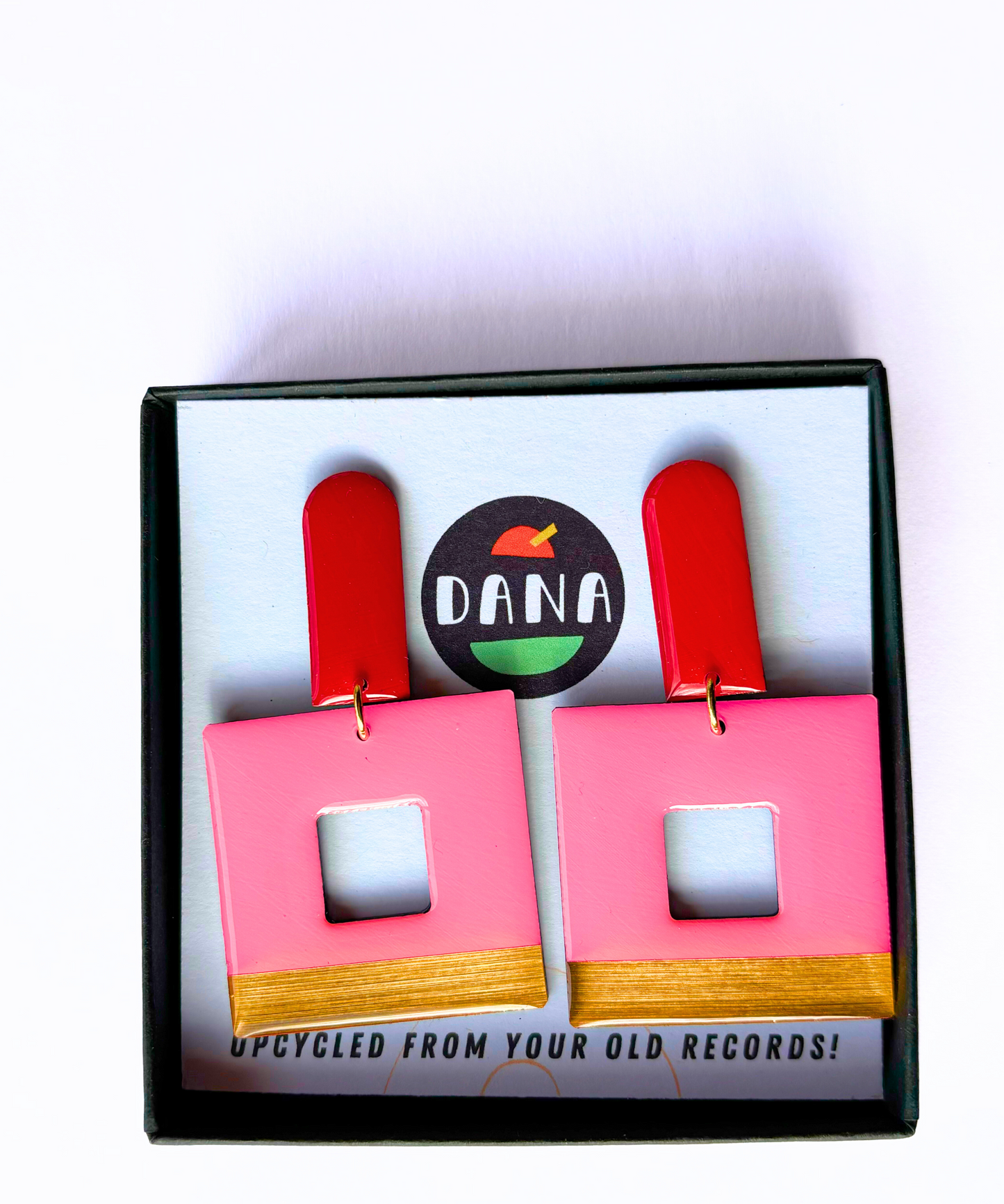 A pair of eco friendly geometric statement earrings with red long arch posts, pink, and gold colours with a bold design, displayed on a card with the DANA branding crafted in Ireland from recycled vinyl record. 