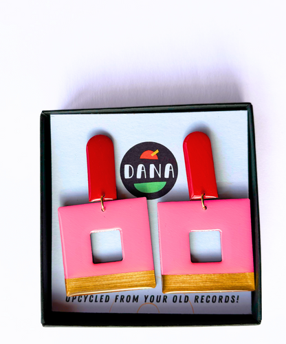 A pair of eco friendly geometric statement earrings with red long arch posts, pink, and gold colours with a bold design, displayed on a card with the DANA branding crafted in Ireland from recycled vinyl record. 
