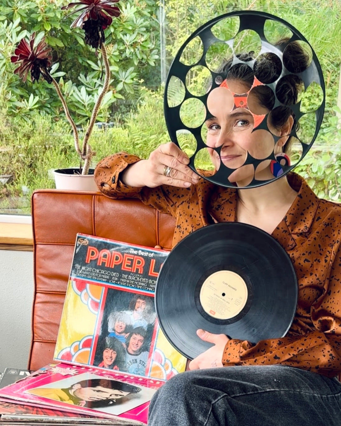 Anne-Louise Laflamme is the designer-maker behind Irish brand DANA Jewellery, defined by the creative reuse of recycled vinyl record. On this image she holds a vinyl record while sitting on an orange chair with vinyl records in sleeves on it and peeking through, with a light smile, a cut out vinyl record.